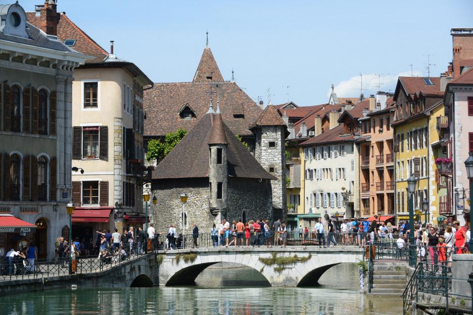 People walk across a stone bridge over a canal in Annecy, France, with historic buildings lining the banks.