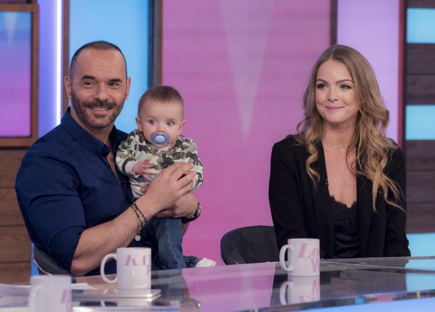 Michael Greco, holding his son Gianluca, and Helen Harris smiling during a television show.