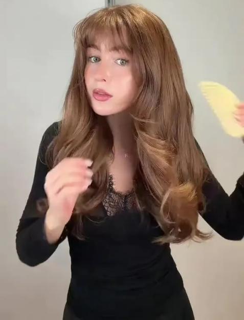 A woman with long brown hair and bangs, wearing a black lace-trimmed top, brushes her hair with a yellow comb.