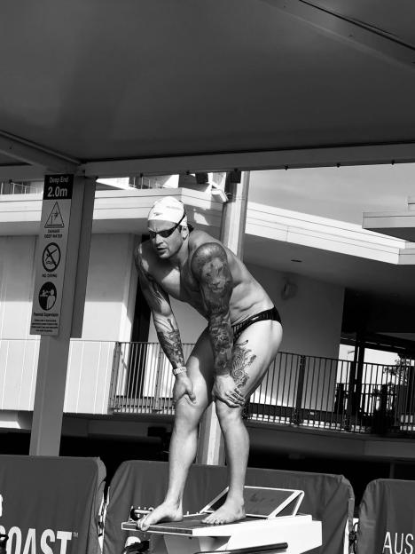 Adam Peaty stands on a swimming block, leaning forward with hands on knees.