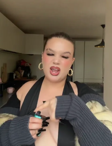 A woman with dark hair, a black top, and a fluffy jacket is pointing at a small green device.