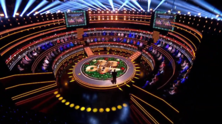 A game show set with a circular stage in the center, a host, and audience members seated in tiered rows.