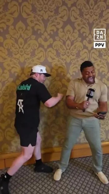 Canelo Alvarez playfully punches a reporter holding a microphone and looking at his phone against a patterned wallpaper background.