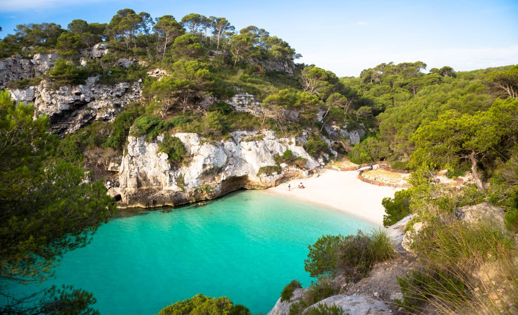 A secluded beach in Menorca, Spain, with turquoise waters, white sand, and lush green trees on rocky cliffs.