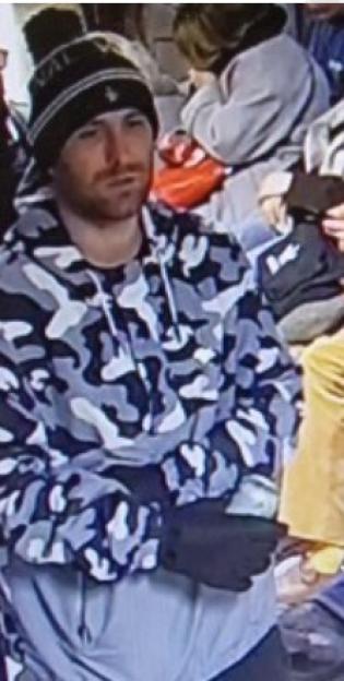 A man with a beard and dark beanie with a pompom, wearing a grey camouflage hoodie and black gloves.