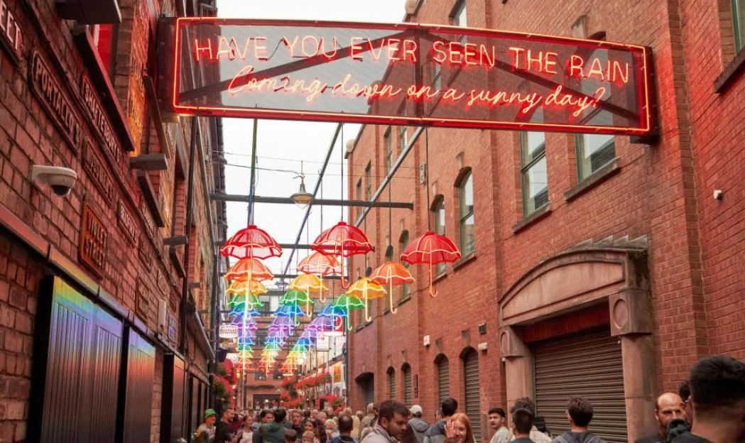 A narrow alleyway between red brick buildings, with a large glowing neon sign overhead that reads "HAVE YOU EVER SEEN THE RAIN Coming down on a sunny day?". Below the sign, rows of colorful umbrellas hang suspended above a crowd of people.