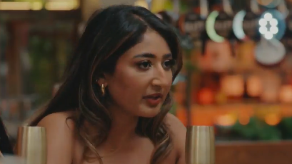 Julia-Ruth with long brown hair, wearing a strapless top and hoop earrings, speaking at a table.