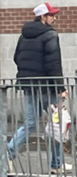 Man in a dark puffer jacket, blue jeans, red and white cap, with dark facial hair, holding a bag.