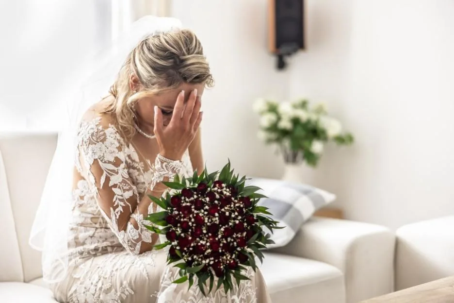A sad bride in a white wedding dress holds her hand to her face in regret while holding a bouquet of red roses.