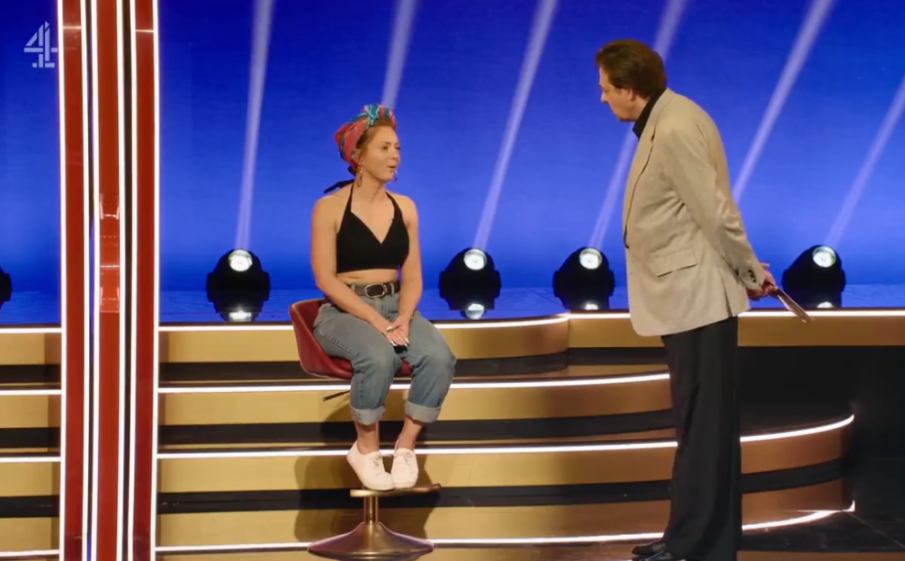 A woman in casual attire sits on a stool opposite a man in a light suit holding a golden spatula, both on a game show stage with blue and gold lighting.