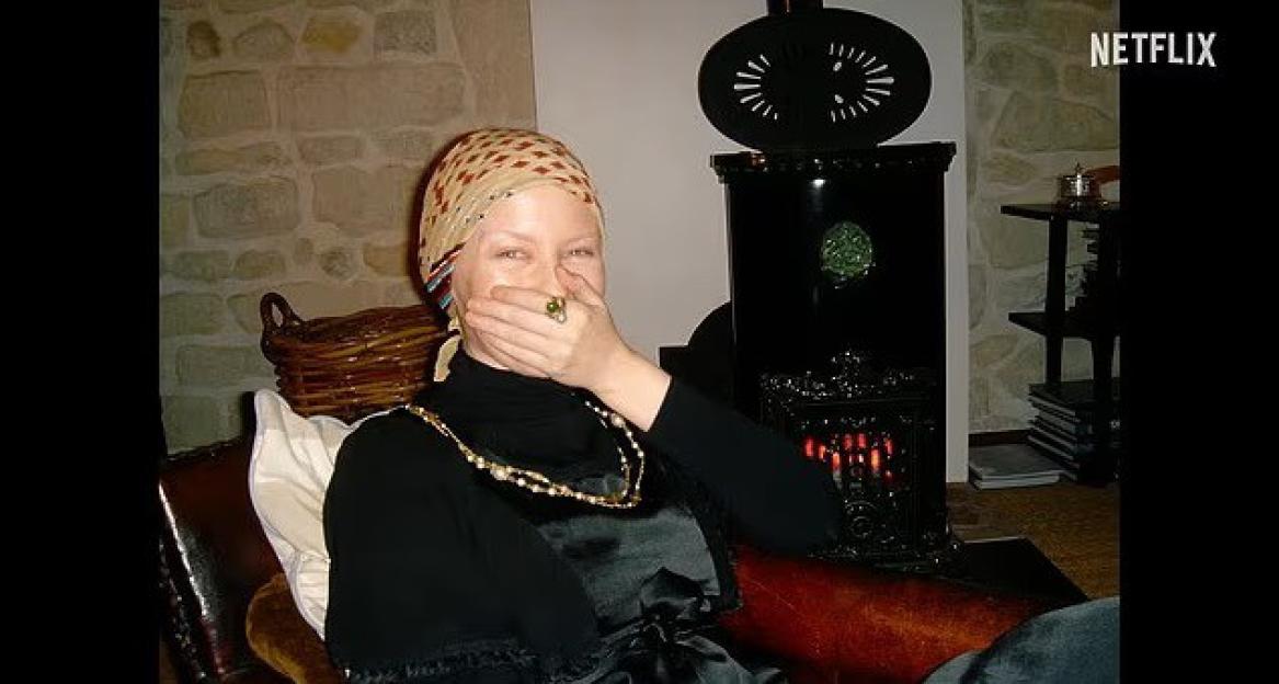A person with a patterned headscarf and a ring on their finger covers their mouth while sitting indoors.