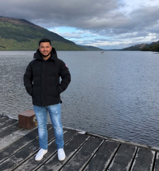Najib Maghsodi standing on a wooden pier next to a lake with mountains in the background.