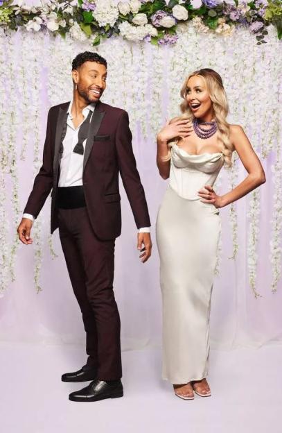 A man in a tuxedo and a woman in a white dress, with a floral backdrop.