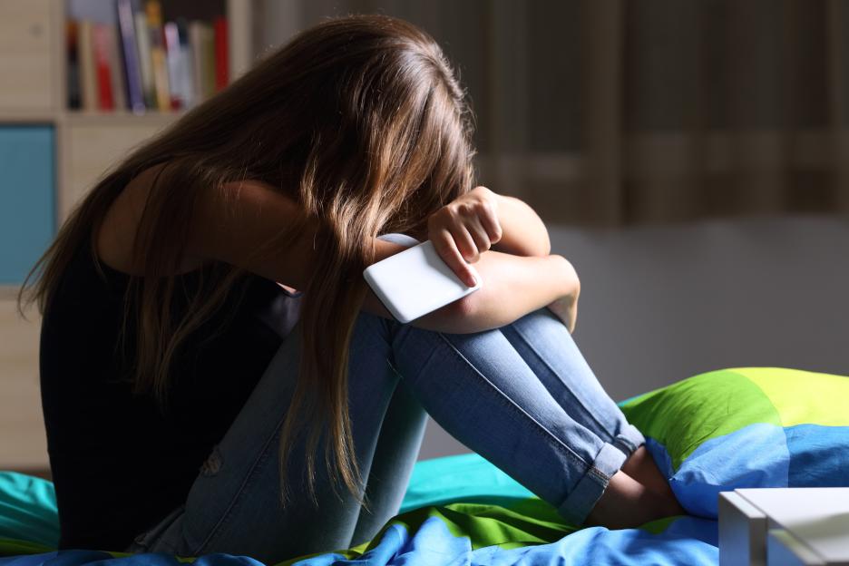 A sad teen sits on her bed in a dark room with her face buried in her arms, holding a phone.