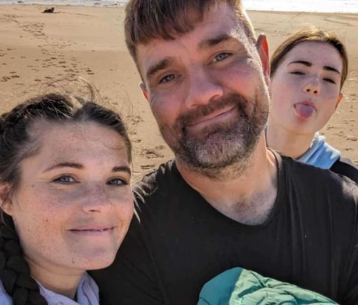 A family selfie on a beach showing a woman with braided hair, a bearded man, and a young girl sticking out her tongue in the background.