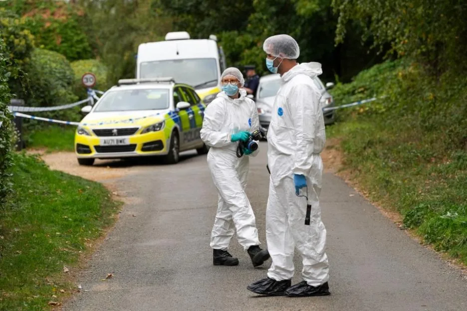 Forensic investigators in white protective suits stand at a crime scene with police cars in the background.