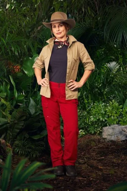 Coleen Rooney in a tan shirt, red trousers, and a cowboy hat standing in a jungle setting.