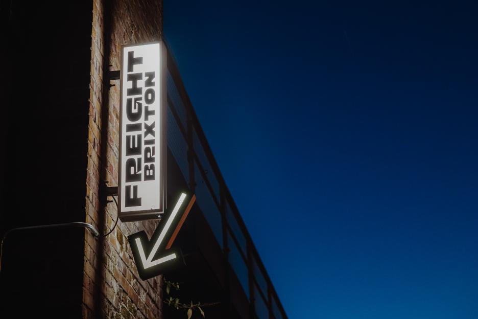 An illuminated sign for "Freight Brixton" with an arrow pointing down, attached to a brick wall.