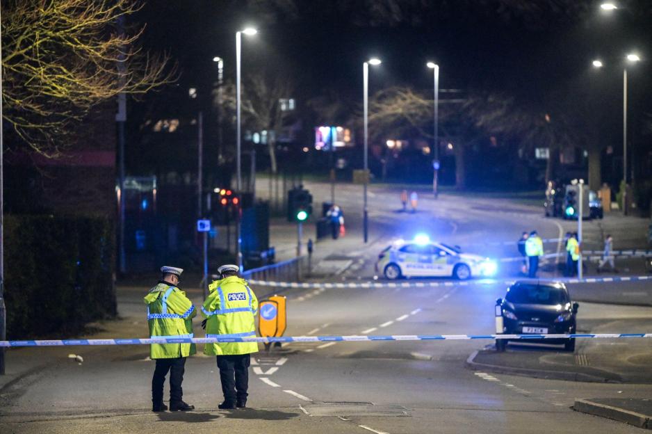 Police officers standing in a cordoned-off street at night with a police car in the background.