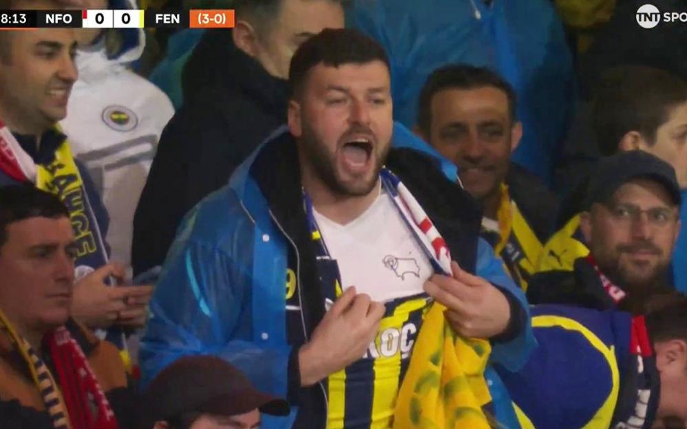 A man in a blue coat pulls open his shirt to reveal a white t-shirt with a ram logo, celebrating with his mouth open in a crowded stadium.