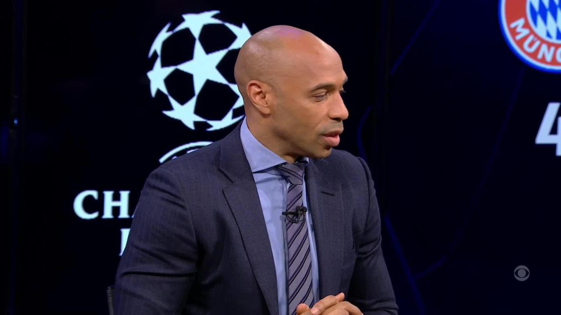 Thierry Henry in a suit and tie, speaking against a background featuring the UEFA Champions League logo.