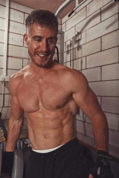 A shirtless, muscular man smiling in a gym setting.