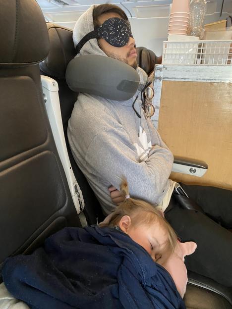 Andy sleeps with a sleep mask and neck pillow in an airplane seat, while his daughter Malva sleeps on his lap.