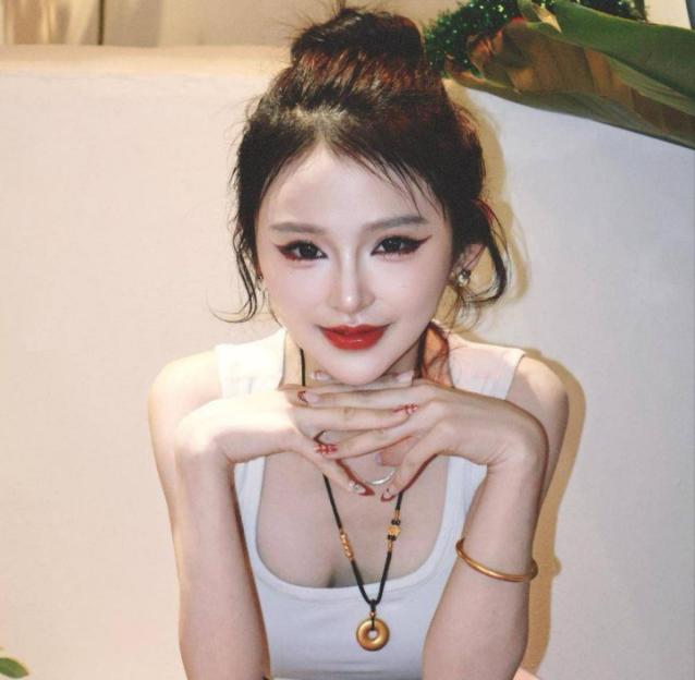 A young woman with dark hair in a bun, red lipstick, and winged eyeliner smiles at the camera, wearing a white tank top and a necklace with a round pendant.