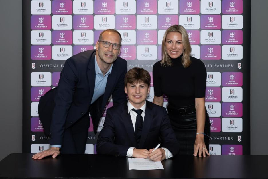 A young man in a suit signs a document, flanked by a man and a woman.