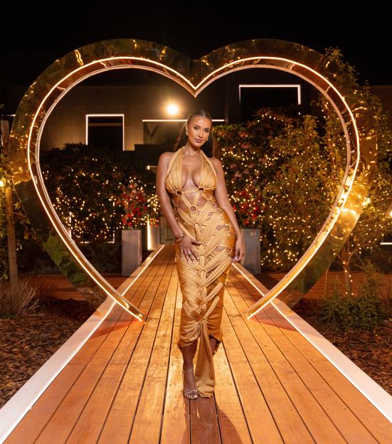 Maya Jama posing in a gold dress with a large heart-shaped arch behind her for "Love Island: All Stars" TV Show in South Africa.