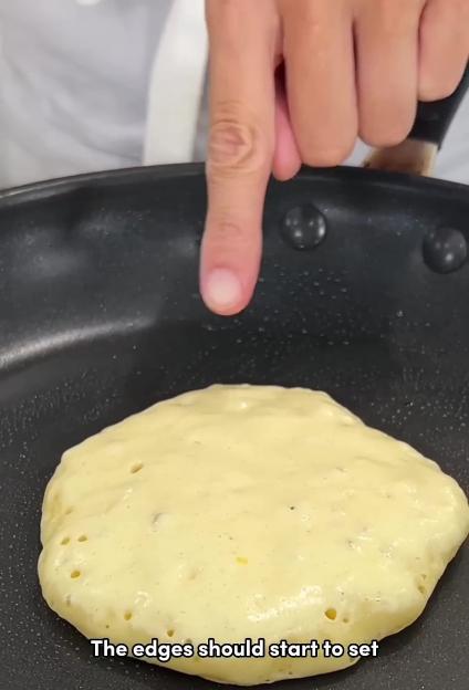 A hand points at the setting edge of a pancake cooking in a frying pan.