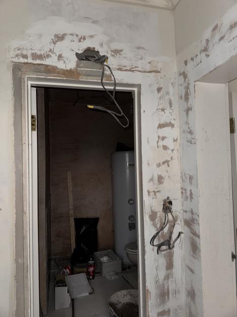 An unfinished home construction with exposed plaster, wires, and a water heater visible through an open doorway.
