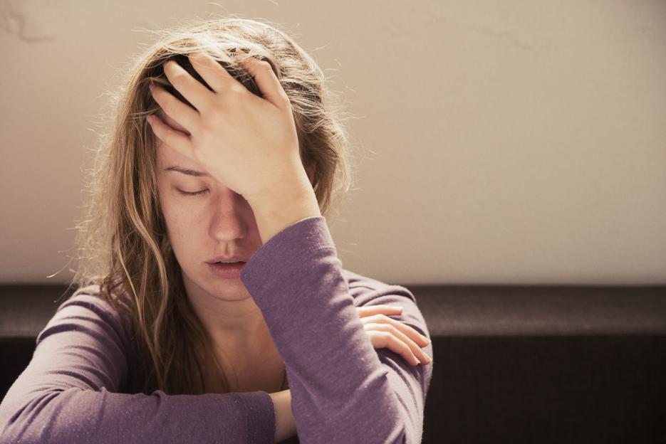 A woman in a purple long-sleeved shirt covers her face with her hand, grimacing in pain from stress or a headache.