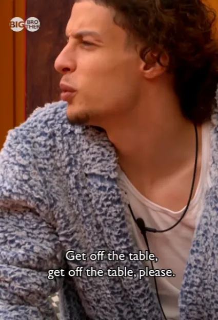 A Big Brother housemate in a blue textured jacket looks away with a serious expression, with subtitles reading, "Get off the table, get off the table, please."