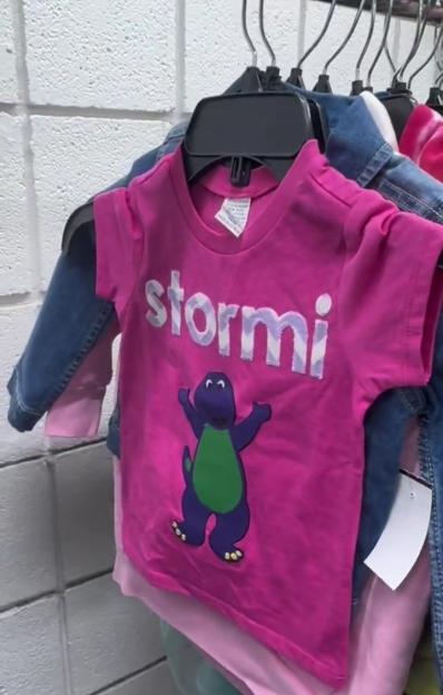 A pink children's t-shirt with a purple Barney dinosaur graphic and the name "Stormi" above it, hanging on a rack with other clothing.
