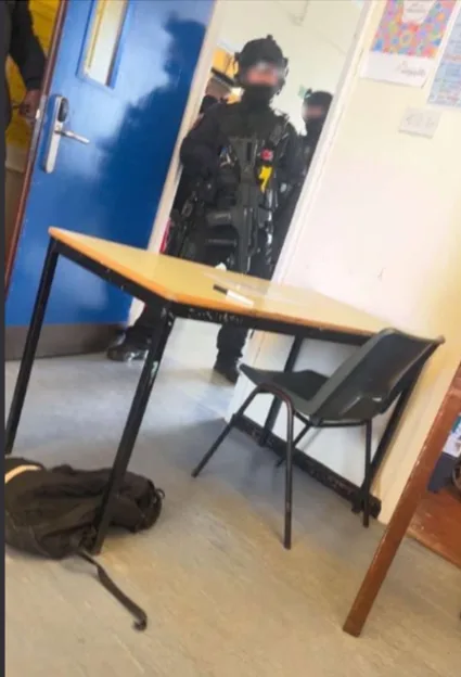Armed police officer in tactical gear standing in a school doorway behind a desk.