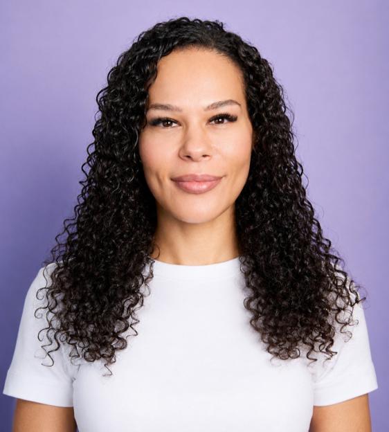 A person with clear skin, curly dark hair, and a white shirt smiling faintly against a purple background.