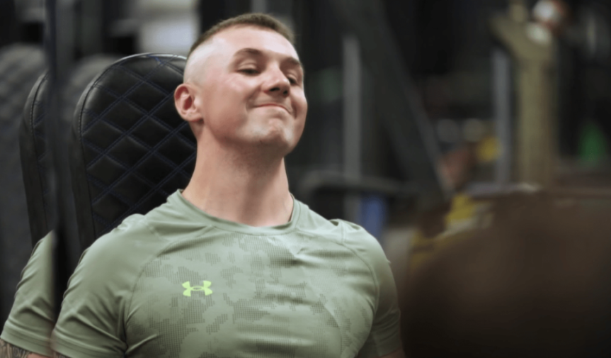 A young man with a buzz cut and green athletic shirt smiles while leaning back on a black quilted seat.