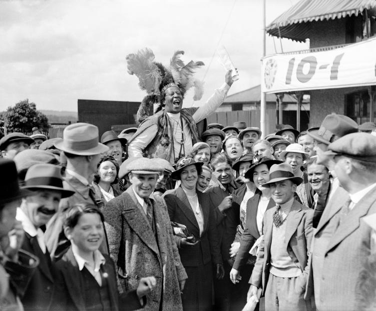 Prince Monolulu in a feathered headdress and beaded vest, enthusiastically addressing a cheering crowd at the Epsom Derby.