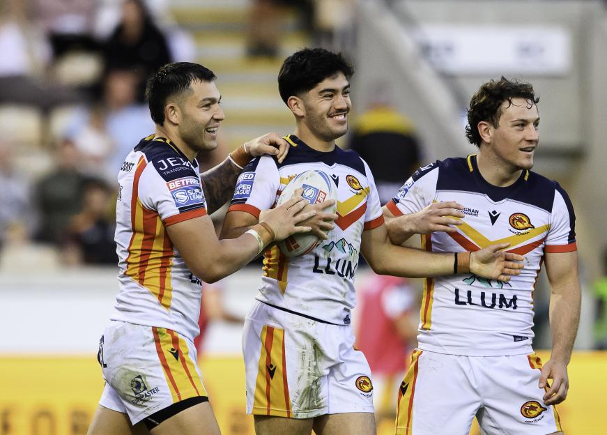 Three smiling rugby players from Catalans Dragons in white, red, and yellow uniforms, two with their arms around the third who is holding a rugby ball.