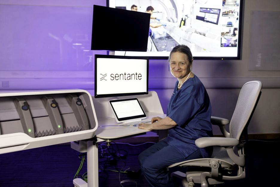 Professor Iris Grunwald in blue scrubs sitting at a robotic surgical system, with "sentante" on the monitor and a screen in the background showing two surgeons and a patient.