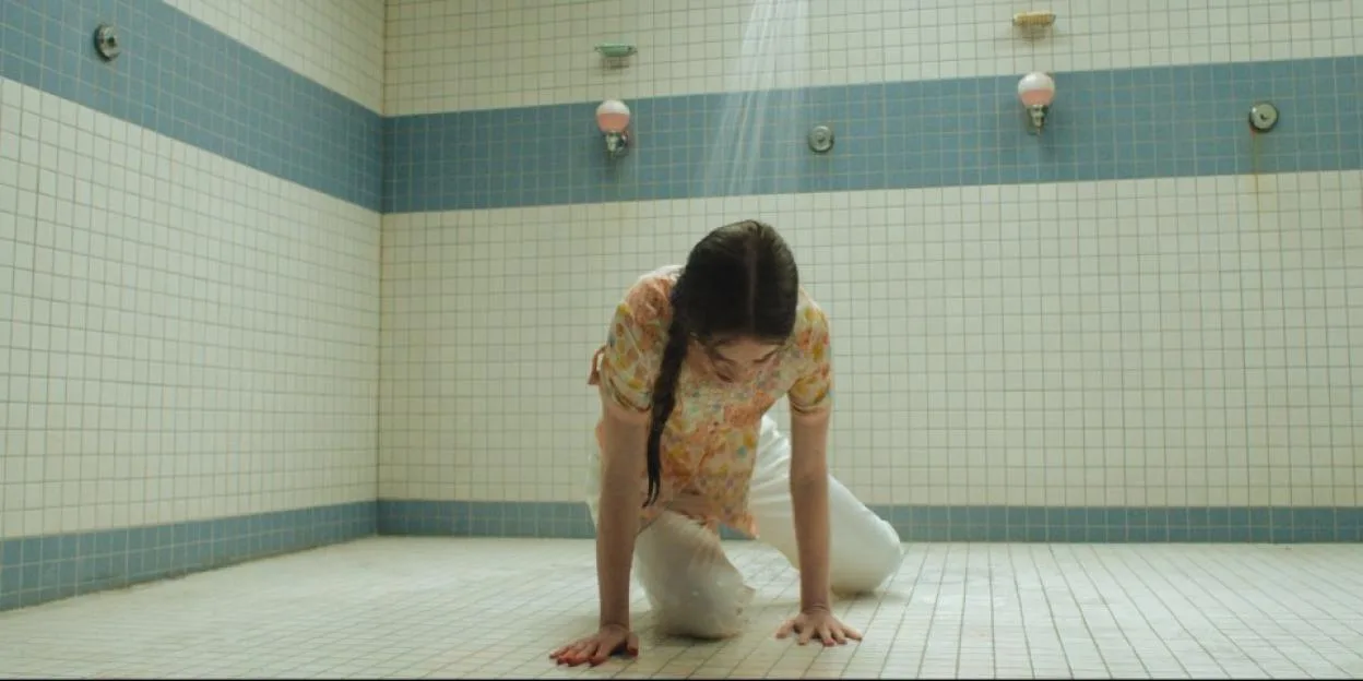 Summer H. Howell kneeling on the tiled floor of a communal shower while water runs down from above.