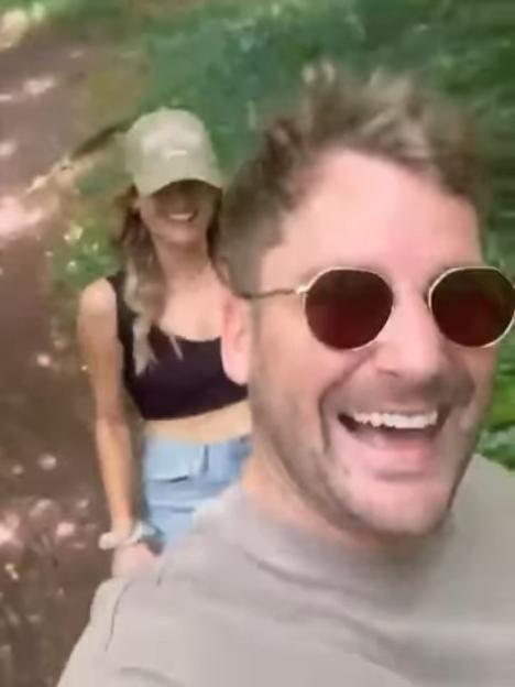 A man wearing sunglasses smiling and a woman in a cap walking behind him.