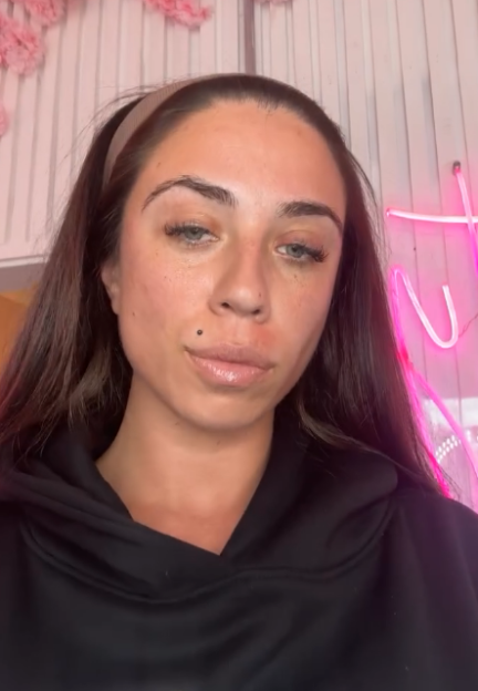 A woman in a black hoodie with dark hair and a headband looks at the camera, with a pink neon light visible behind her.