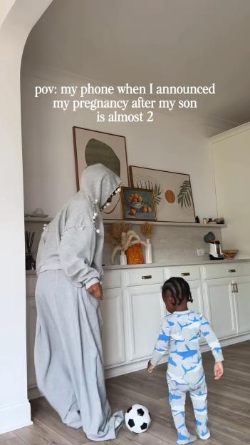 A woman in a gray hoodie with pearl details, standing in a kitchen with a young boy in shark pajamas, as text reads, "pov: my phone when I announced my pregnancy after my son is almost 2."