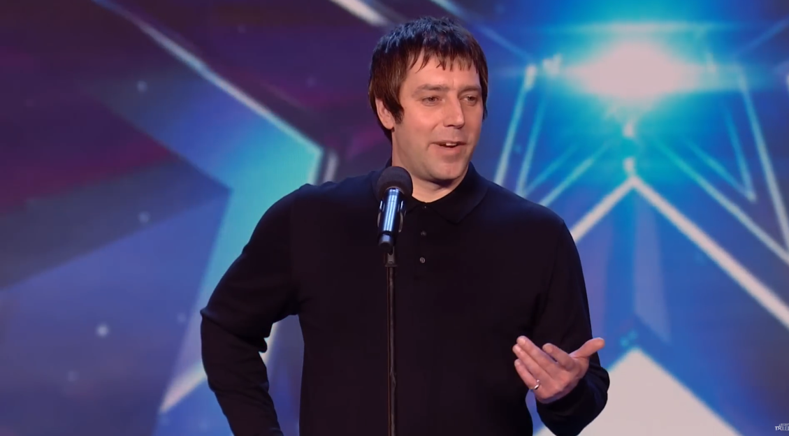 A man in a black long-sleeved polo shirt speaks into a microphone on a stage with blue lighting.