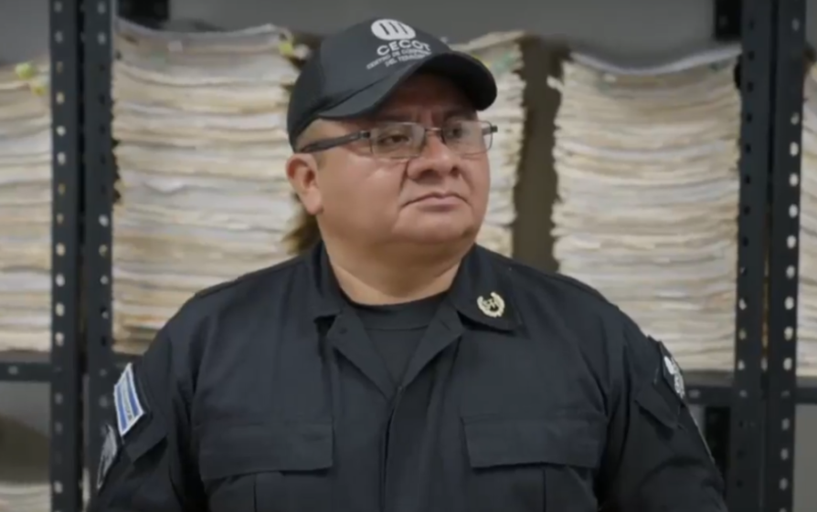 A man in uniform wearing glasses and a cap, with large stacks of paper in the background.
