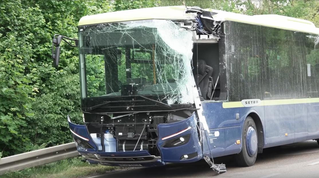 A bus with a severely damaged front, including a shattered windshield and missing bumper pieces.