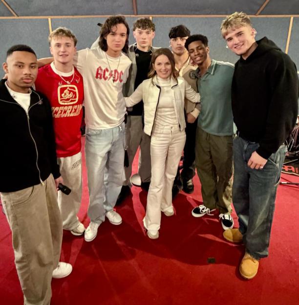 Geri and December 10, a group of seven young men and one woman, pose together in a room with a red carpet and acoustic foam walls.