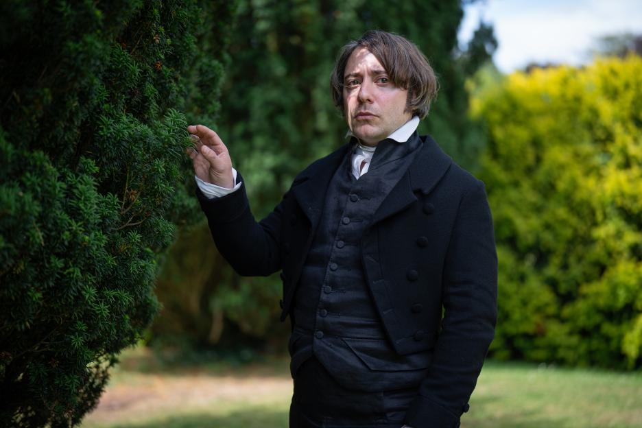 Mr Collins (Ryan Sampson) in period attire, touching a green bush.